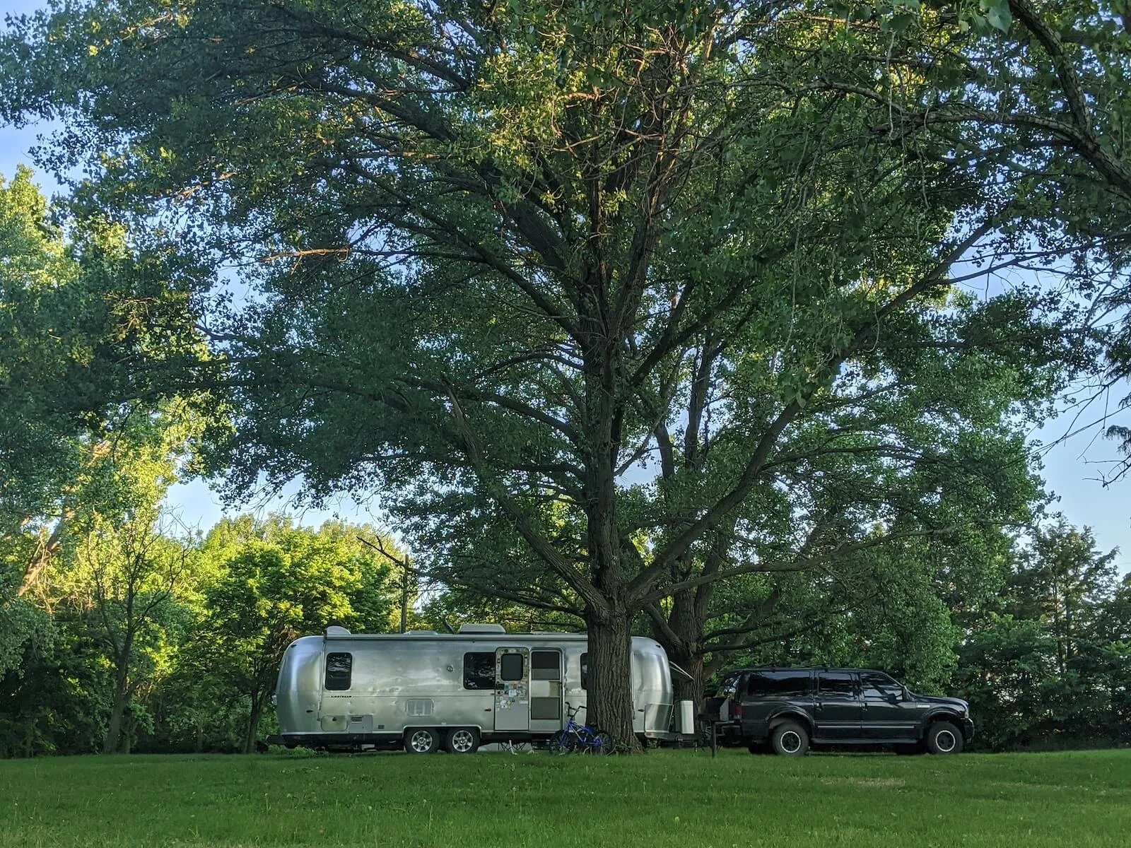 large truck and airstream rv parking underneath large tree with grassy field in the foreground travel nursing in an RV