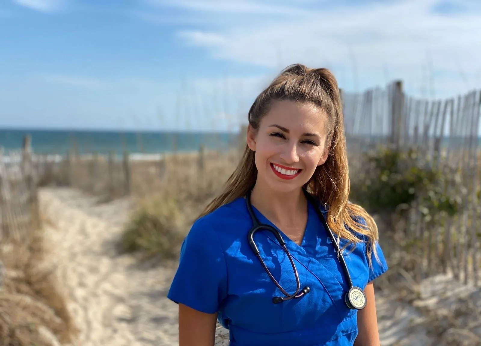 A woman smiles on the beach wearing blue Cherokee nurse scrubs.