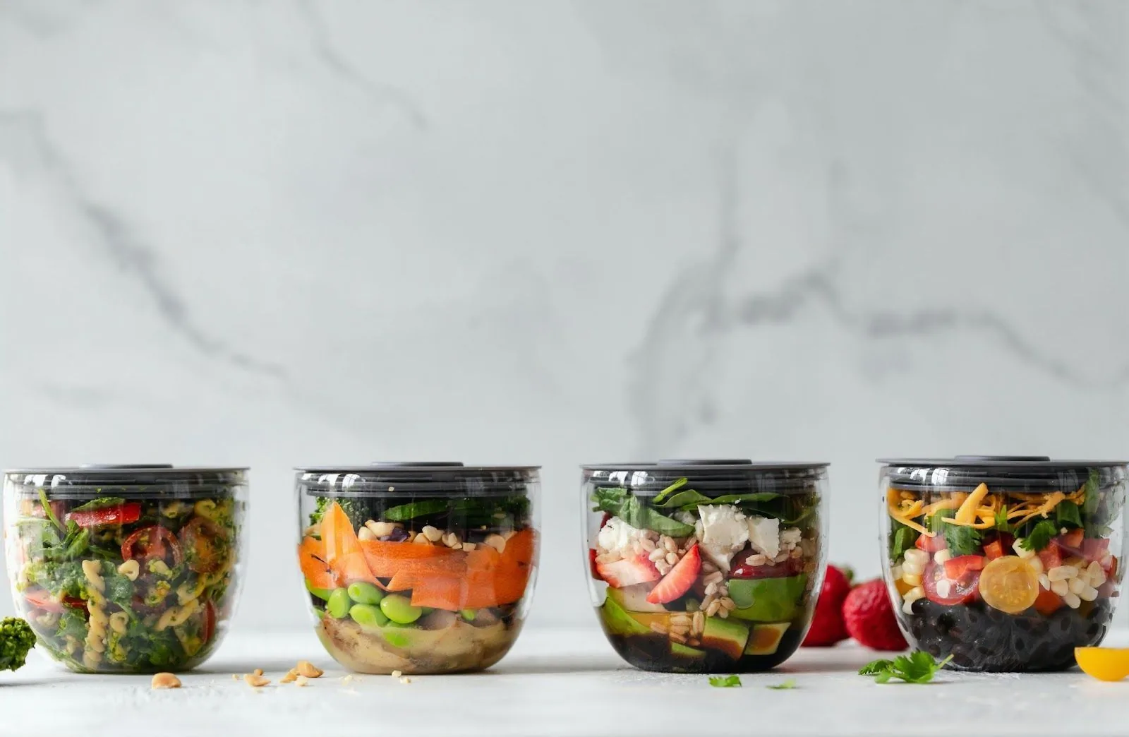 Four clear jars filled with a variety of colorful fresh vegetables, fruits, and grains on a light marble background, representing healthy snack options for the holidays.