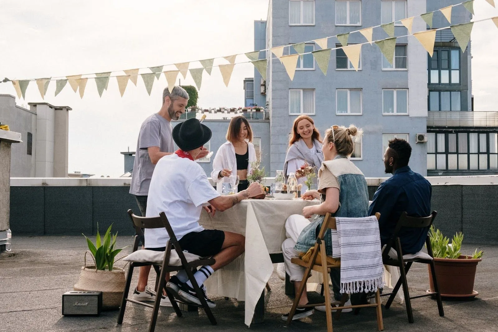 A diverse group of people enjoying a rooftop party with festive bunting, engaged in conversation around a table with drinks and snacks, embodying the joy of socializing with colleagues in a new city during the holidays.