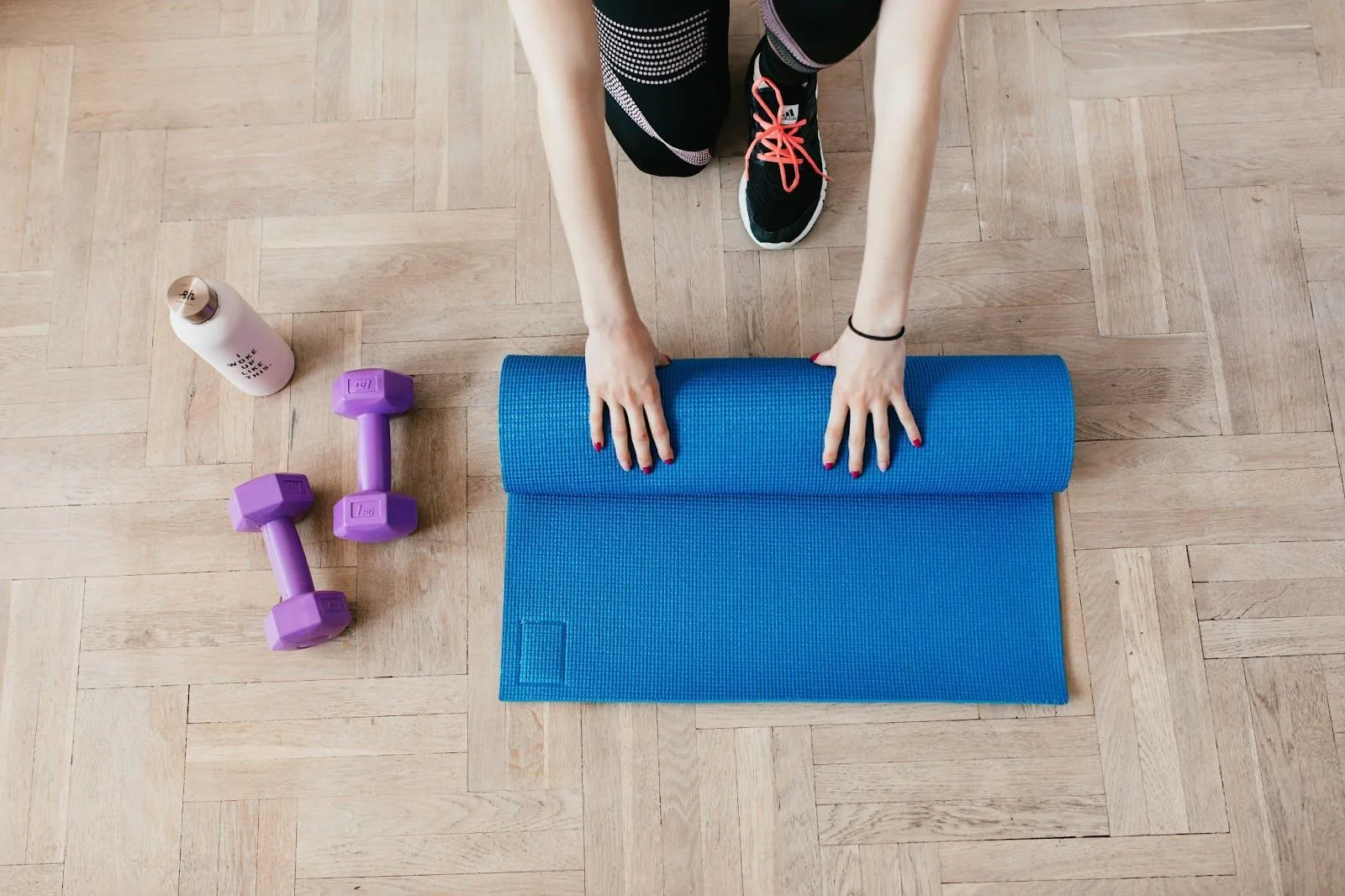 A top-down view of a person kneeling on a wooden floor, rolling out a vibrant blue yoga mat next to purple dumbbells and a white water bottle, symbolizing the convenience of staying fit with Gympass while traveling.