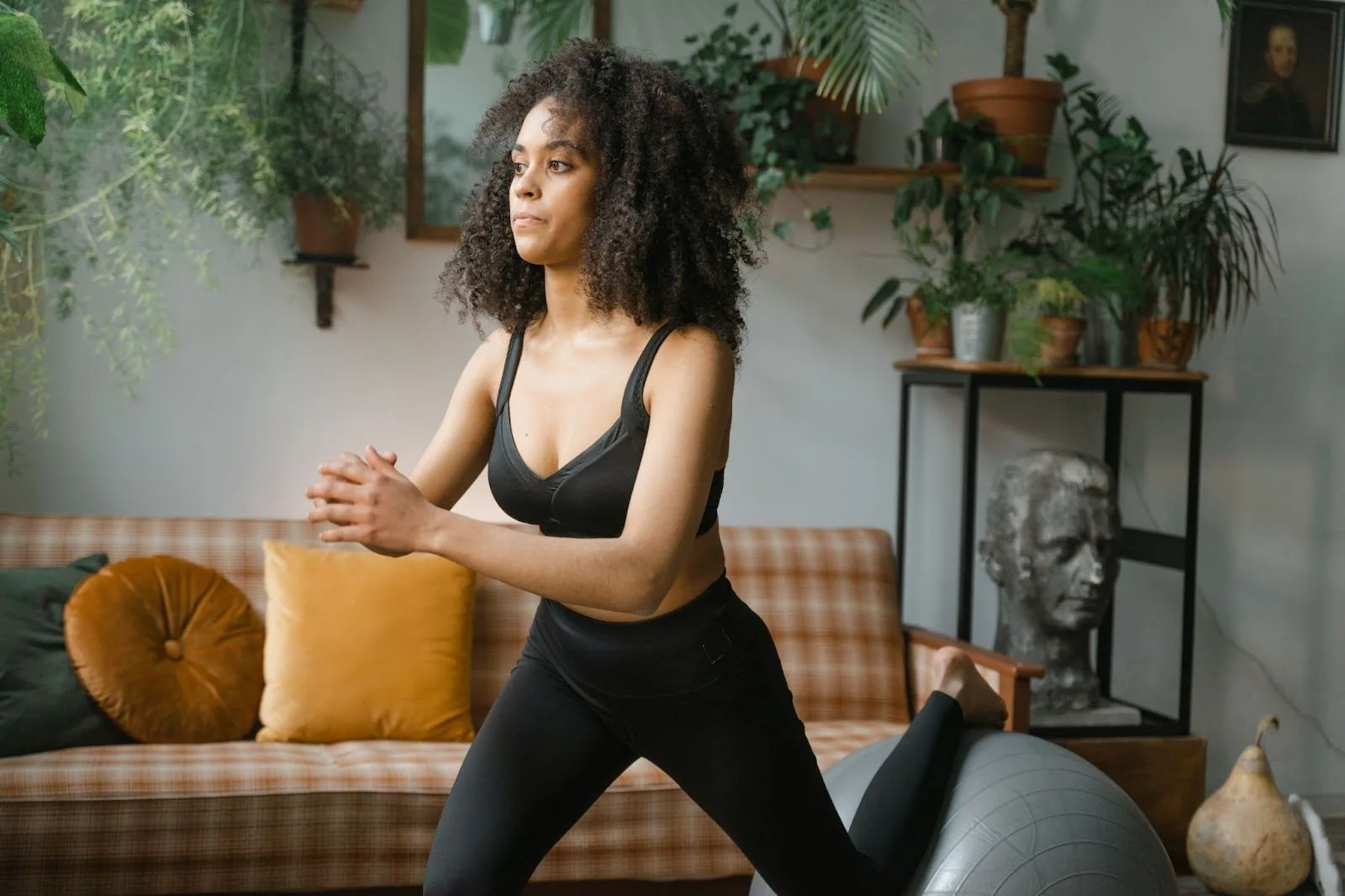 A woman in black activewear performs a lunge on a silver exercise ball in a cozy room filled with plants and natural light, demonstrating an at-home workout setup conducive to fitness while traveling.
