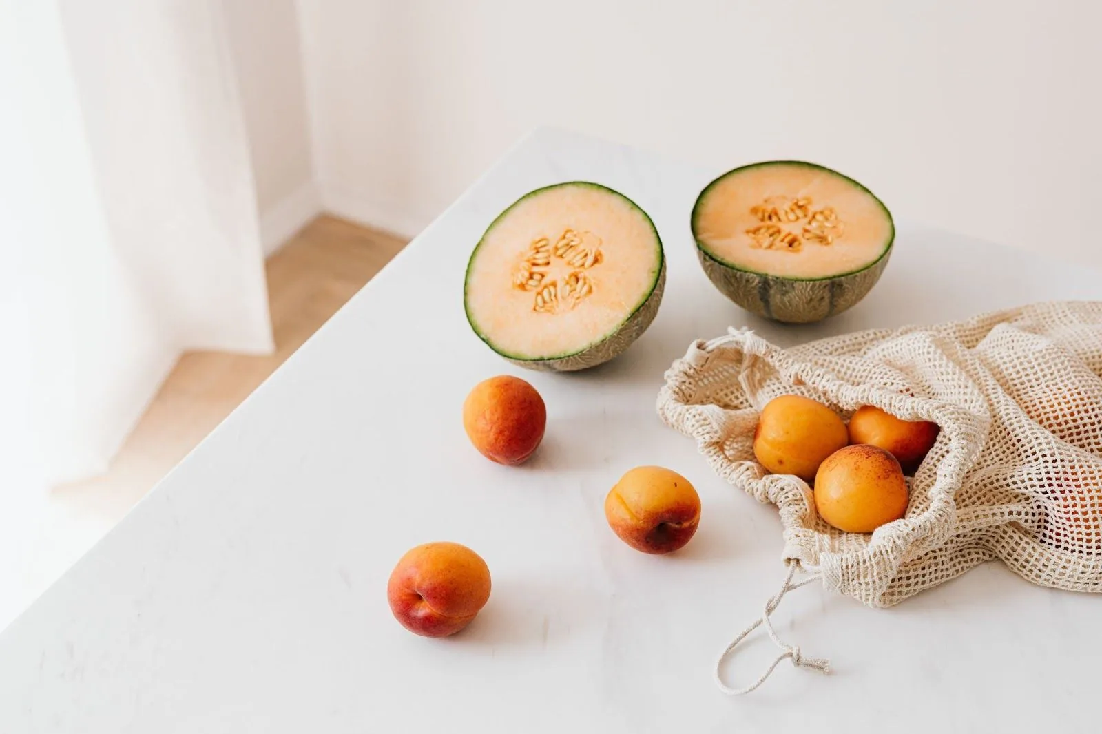 A neatly arranged scene on a white marble countertop showing half of a cantaloupe, a sliced cantaloupe, and apricots spilling from a beige mesh reusable grocery bag, symbolizing healthy eating habits.