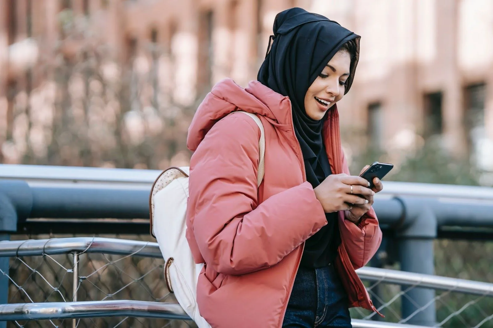 A woman in a pink winter coat and black headscarf smiles while looking at her smartphone, symbolizing the joy of connecting with loved ones through technology during the holidays, even while on the go.