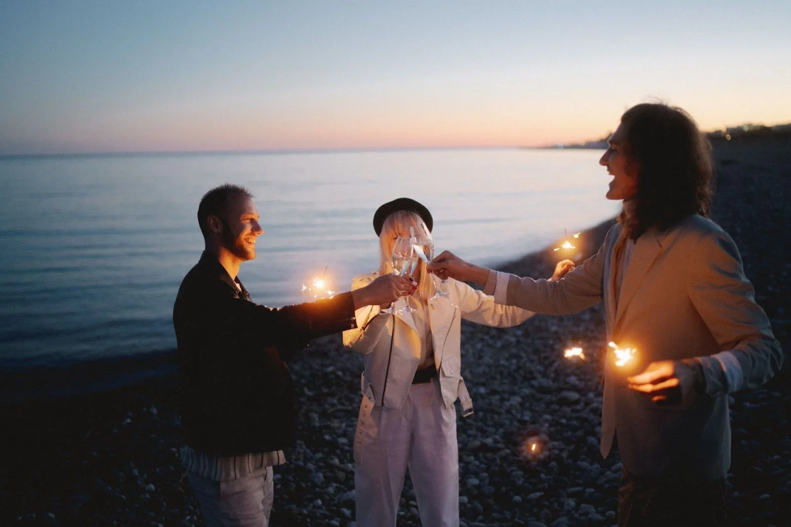 Three people stand on a pebble beach at dusk, toasting with sparkling wine and holding sparklers, exemplifying the warmth of connecting with colleagues and friends during the holidays.