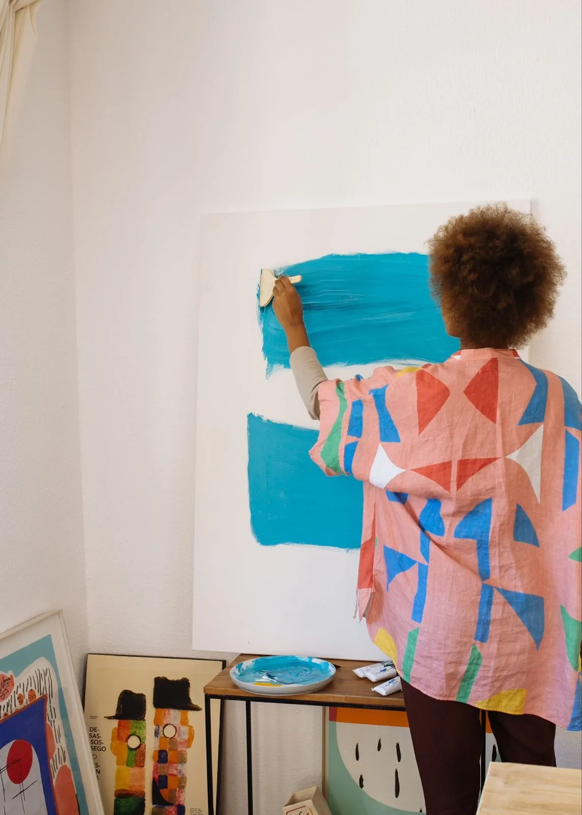 A creative travel nurse with Black descent is painting a vibrant blue stroke on a canvas, wearing a colorful patterned shirt, in a room with art supplies and framed artwork, embodying the joy of a local art class.