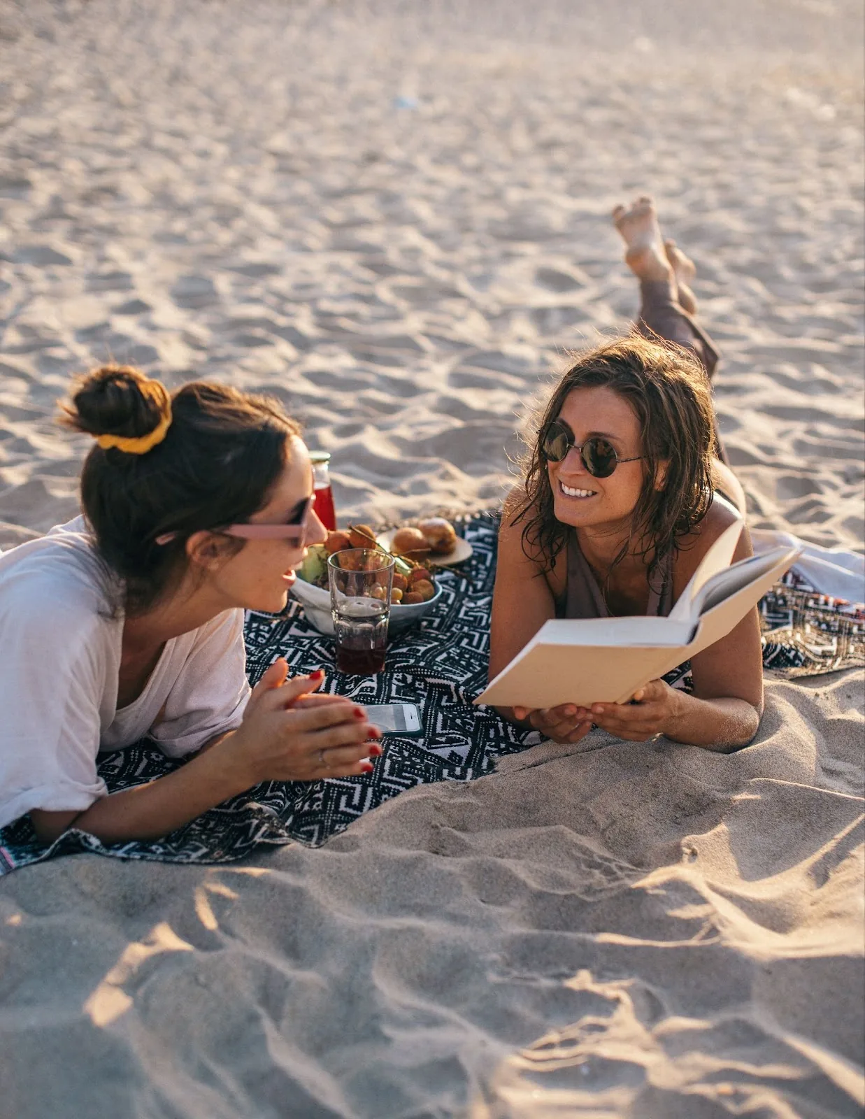 Two women enjoying a relaxing moment on a sandy beach at sunset, with one reading a book and the other smiling, surrounded by a picnic setup, embodying leisure and self-care for a travel nurse.