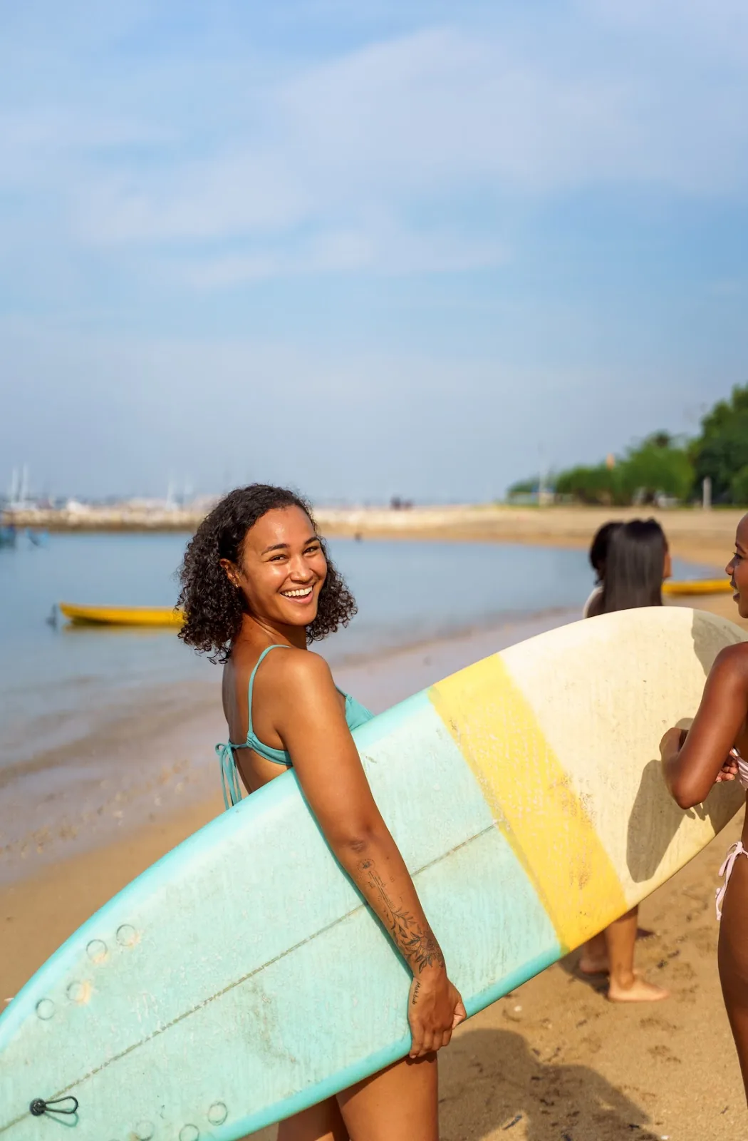 A joyful travel nurse with a mixed-race background is holding a surfboard, smiling widely on a sunny beach with other beachgoers in the background, ready to catch some waves.
