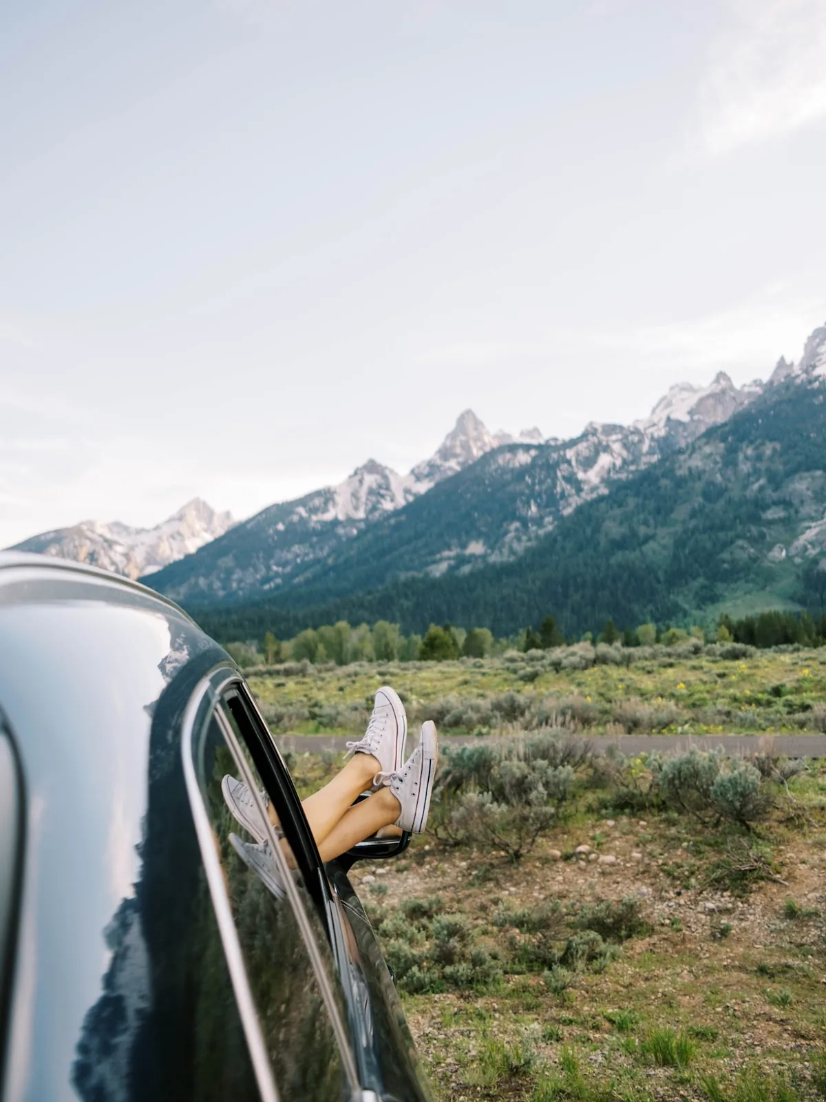 Legs clad in casual footwear protrude from a car window, framing a scenic view of majestic mountains, symbolizing the freedom and exploration a gas gift card can offer a travel nurse