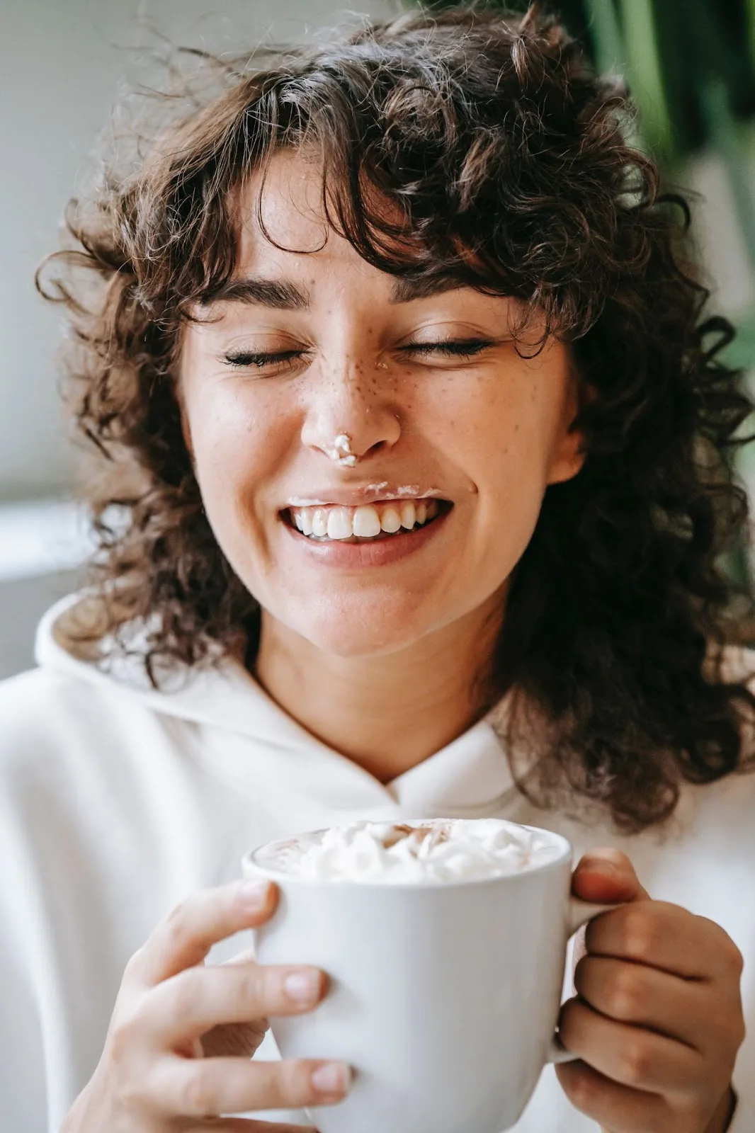 A woman with curly hair and freckles, joyfully closes her eyes, holding a large mug of hot chocolate with whipped cream, hinting at a travel nurse's precious coffee break moments.