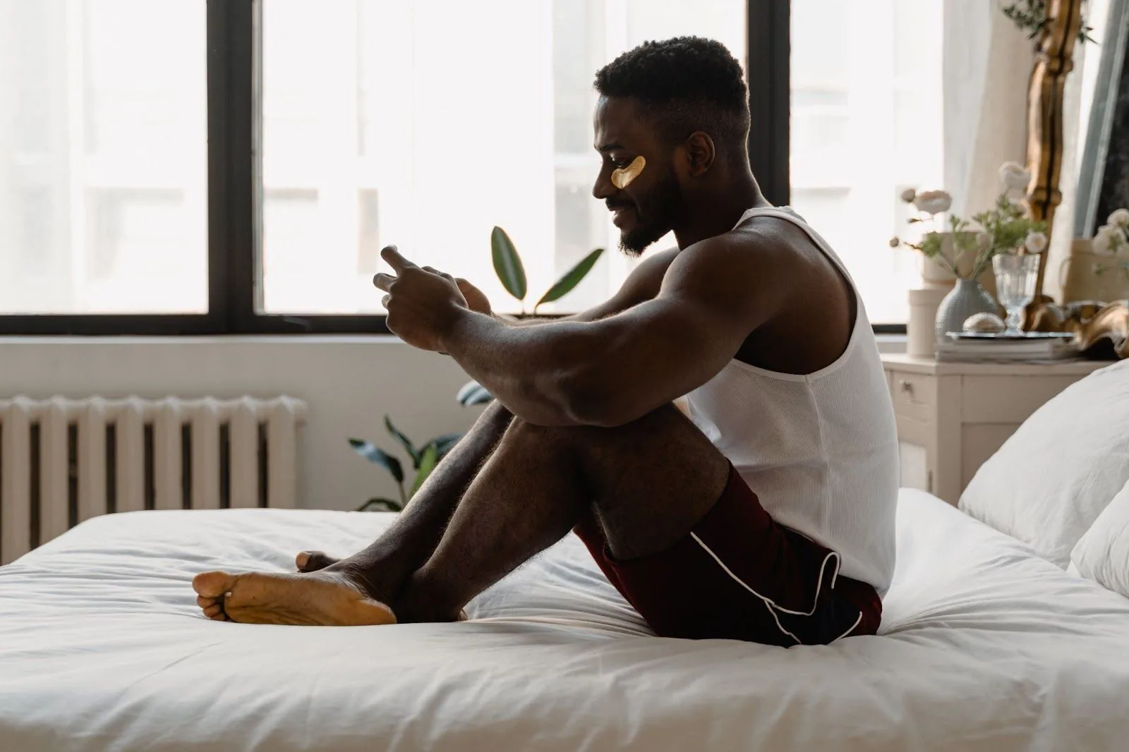 A man sitting on a white bed, engaging in a video call, with golden under-eye patches on, symbolizing self-care and staying connected with loved ones through technology.