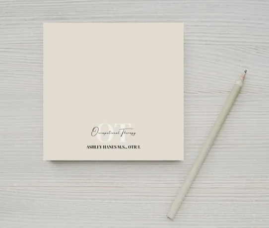 A personalized beige Post-it® note pad lies on a white wooden surface, accompanied by a light taupe pen. The notepad is elegantly printed with 'Occupational Therapy' in cursive and the name 'ASHLEY HAINES M.S., OTR/L' below in a formal font, ready for jotting down reminders and notes.