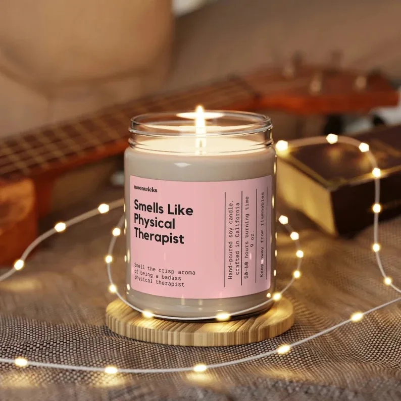 A lit scented candle labeled 'Smells Like Physical Therapist' on a wooden stand, surrounded by warm fairy lights. The candle's label humorously suggests a crisp aroma embodying the spirit of a badass physical therapist, adding a relaxing and affirming atmosphere to any room