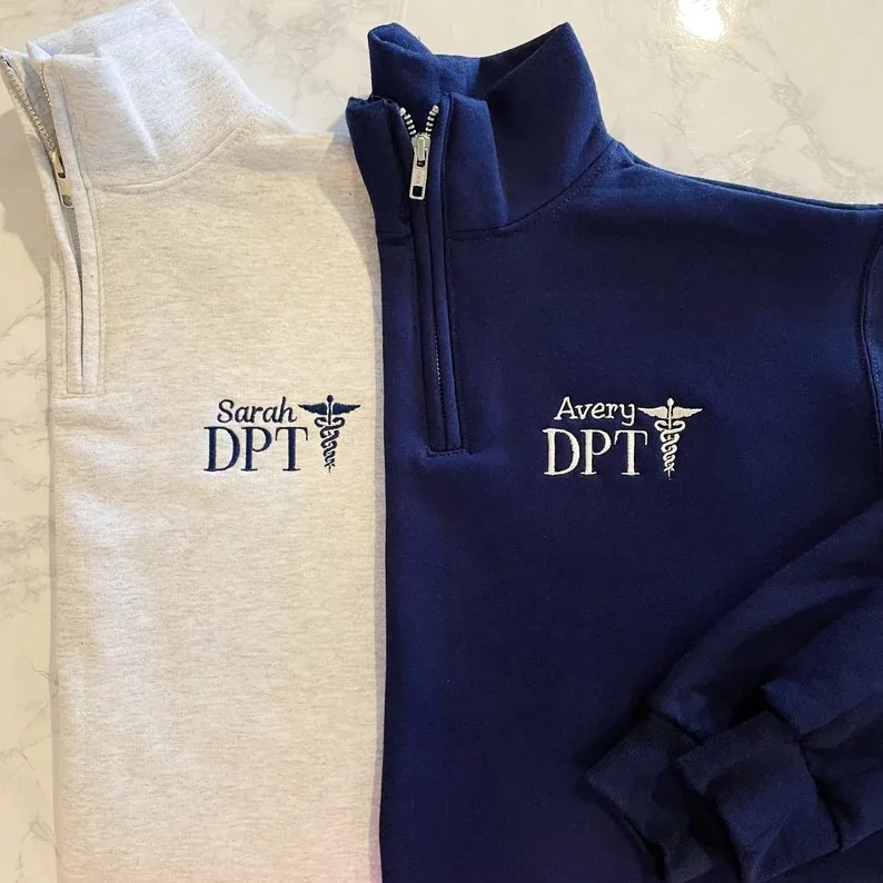 Two quarter zip sweatshirts, one in heather gray and the other in navy blue, each embroidered with a name and 'DPT' credentials. 'Sarah DPT' on the gray and 'Avery DPT' on the navy, showcasing a professional and personal touch for any Doctor of Physical Therapy..