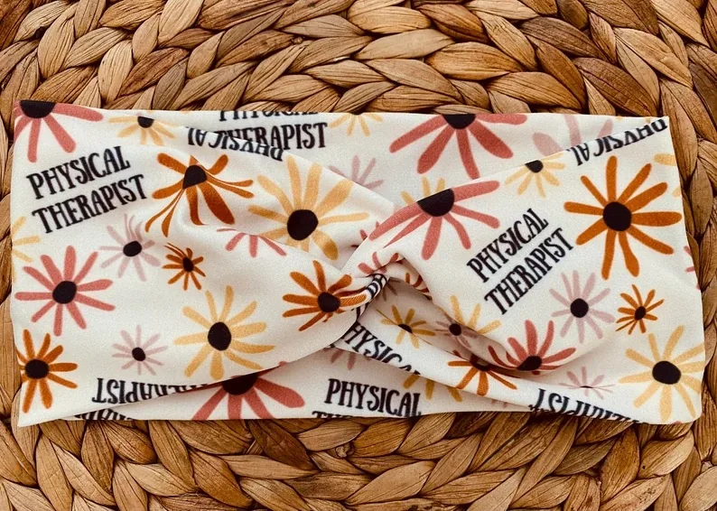 A stylish boho twist headband, adorned with 'Physical Therapist' script interspersed between a pattern of warm-toned daisies, laid out on a woven straw background. Its stretchy material is ideal for busy PTs who want to add a splash of fashion and comfort to their work attire.