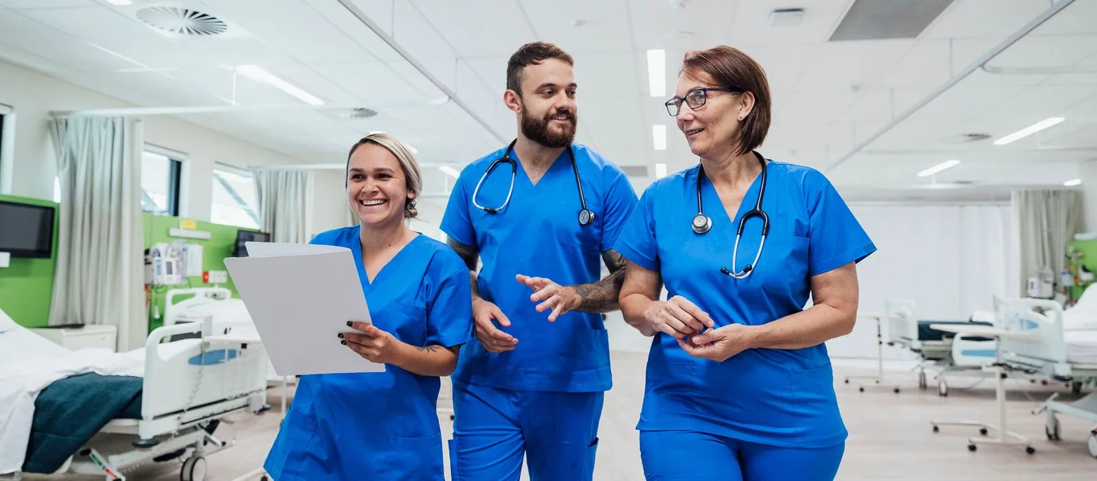 Three smiling healthcare professionals in blue scrubs walking through an ambulatory surgery unit, discussing patient care with a hospital bed and equipment in the background, symbolizing teamwork and patient readiness in a day surgery setting.