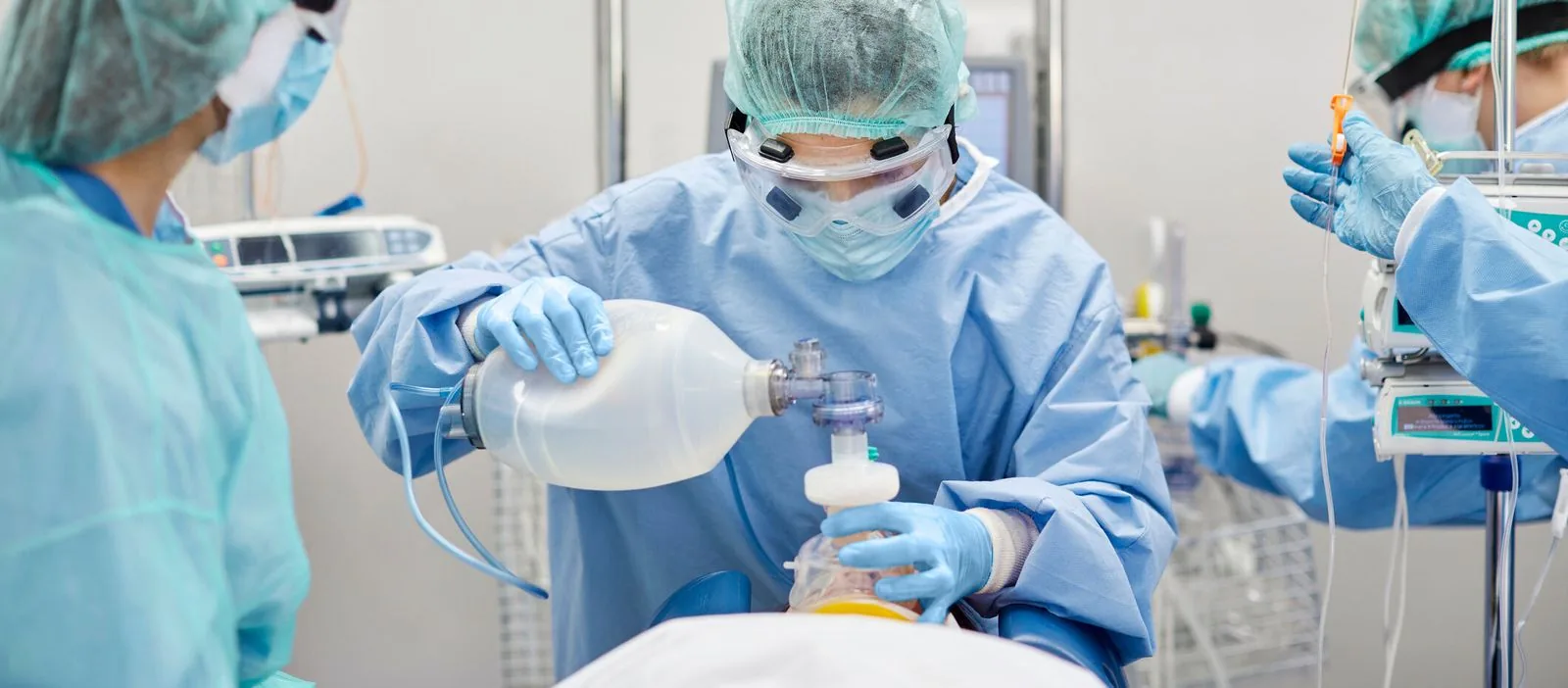 Healthcare professionals in blue scrubs and personal protective equipment intently provide life-sustaining care to a patient in an ICU, with one nurse using a manual resuscitator, illustrating the high level of attention and expertise required in critical care.