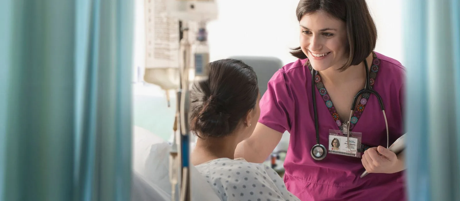 A nurse in a pink scrub with a stethoscope smiles at a patient while holding a clipboard in a bright medical-surgical unit, exemplifying attentive patient care.