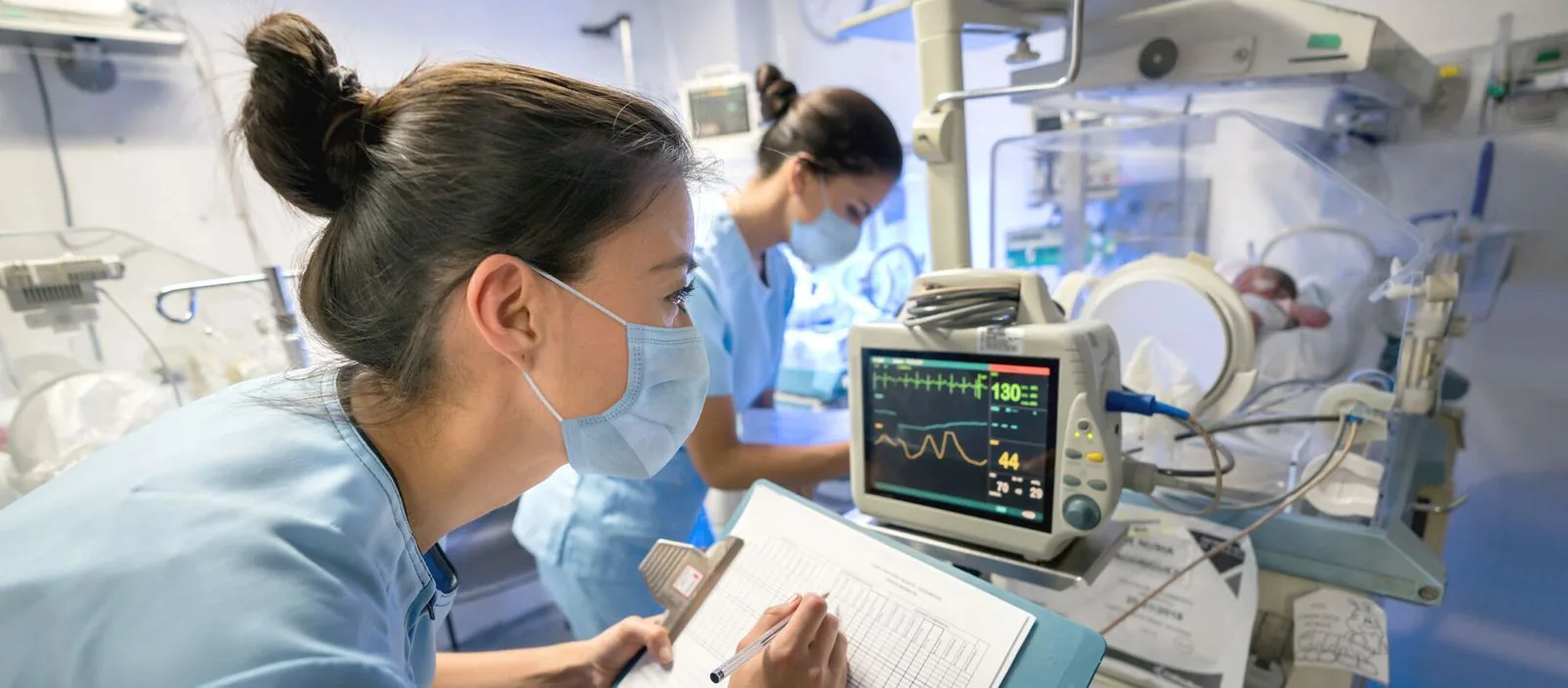 Focused NICU nurse recording data on a clipboard while observing a premature infant in an incubator, with vital signs monitor displaying health parameters in the background.