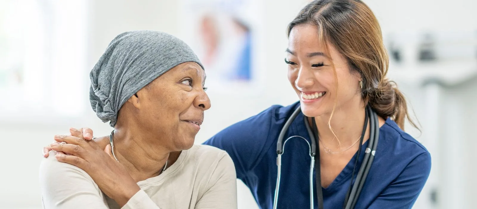 An oncology nurse with a warm smile gently touches the shoulder of a patient in a headscarf, creating a moment of connection in the oncology unit.