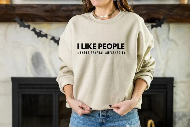A person wearing a beige crewneck sweatshirt featuring the humorous phrase 'I LIKE PEOPLE (under general anesthesia)', available in various colors and styles, perfect for a surgical tech with a sense of humor.