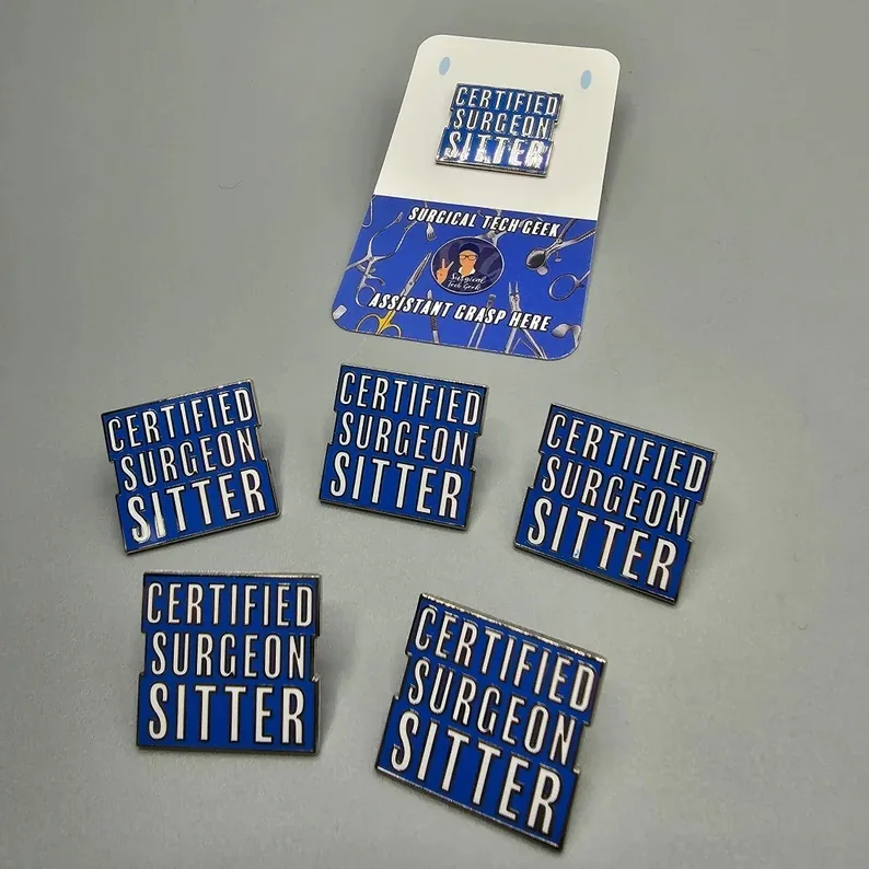 A collection of 'Certified Surgeon Sitter' enamel pins in bold blue and white, designed for surgical techs to wear on scrubs, jackets, backpacks, or attached to badge reels.