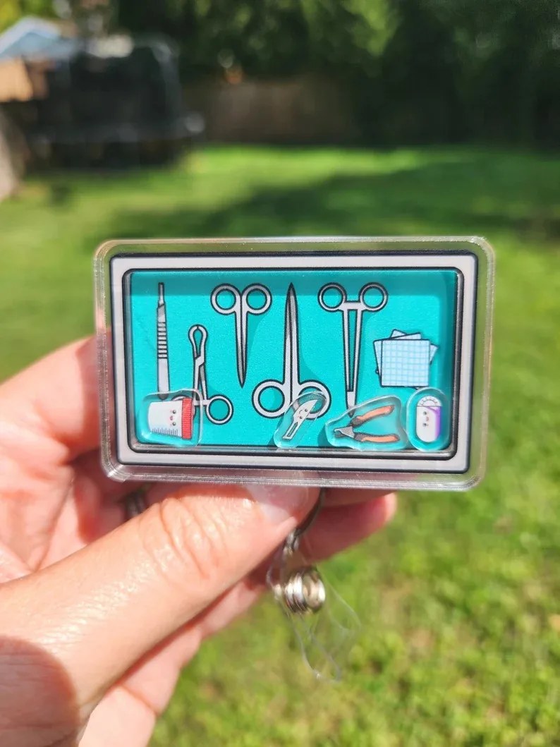 Hand holding a colorful badge reel with a playful array of illustrated surgical instruments on a turquoise background, dangling from a silver clasp, creating a fun and functional accessory for healthcare professionals.