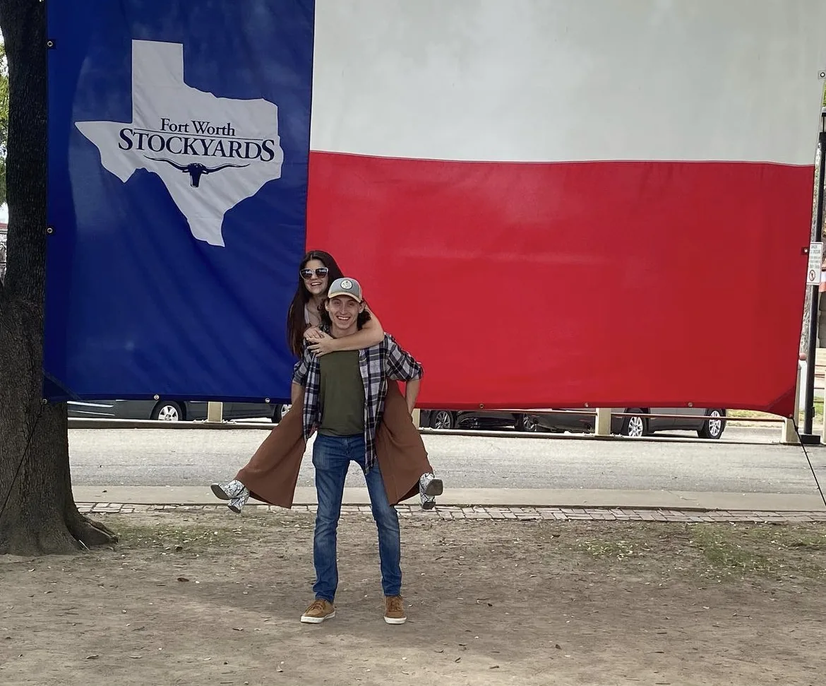 The author piggybacking on her husband in front of the iconic Fort Worth, TX Stockyards flag, celebrating securing her Texas OT licensure.